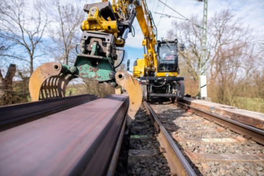 Zeigt einen Bagger, der eine Bahnschiene hebt.